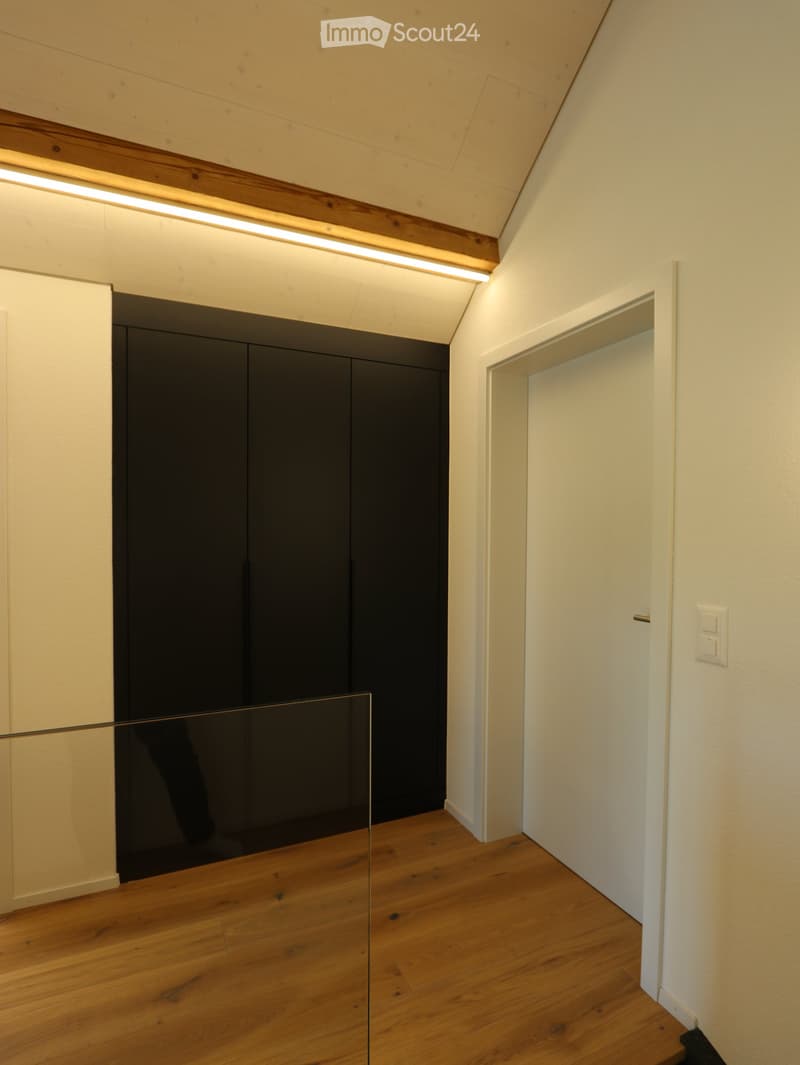 Modern hallway with wooden flooring, white walls, and a sleek black cabinet. Features a glass railing and recessed lighting.