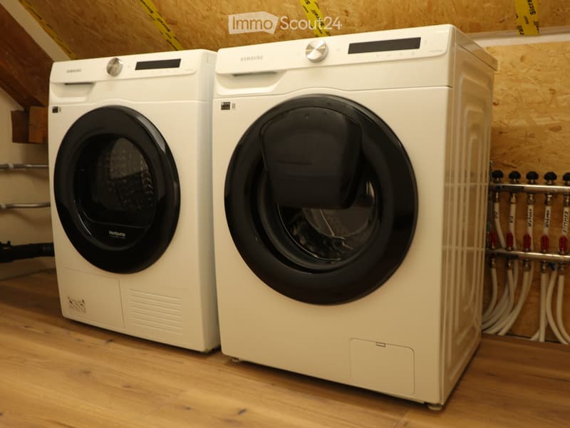 Two white Samsung washing machines and dryers placed side by side on a wooden floor, with exposed wooden beams and a visible heating system in the background.