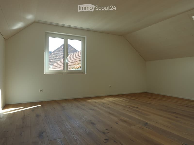 Empty room with wooden flooring and a large window showing a roof view. The walls are painted a light color, and the ceiling has exposed wooden beams.