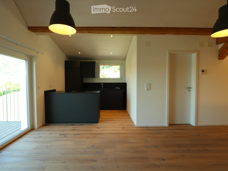 Open-plan space with wooden flooring, exposed wooden beams, modern black kitchen area, large windows, and pendant lights.