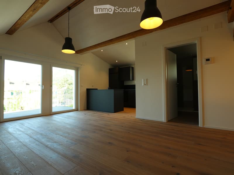 Bright, empty room with wooden flooring, exposed wooden beams, large sliding glass doors leading to an outdoor area, and a small kitchen area visible in the background.