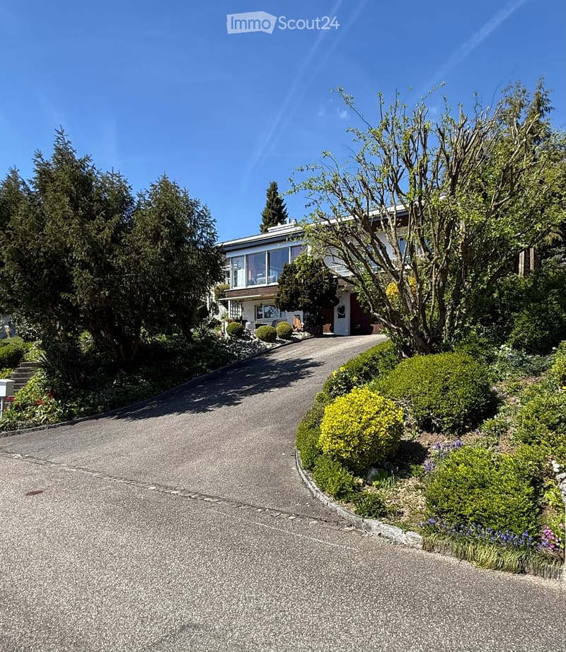 Two-story house with a modern design, surrounded by lush greenery and a well-maintained garden. The house features large windows and a covered entrance.