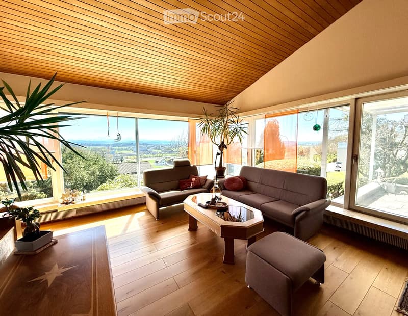 Bright living room with large windows offering a scenic view, featuring a grey sofa, coffee table, ottoman, and decorative plants.