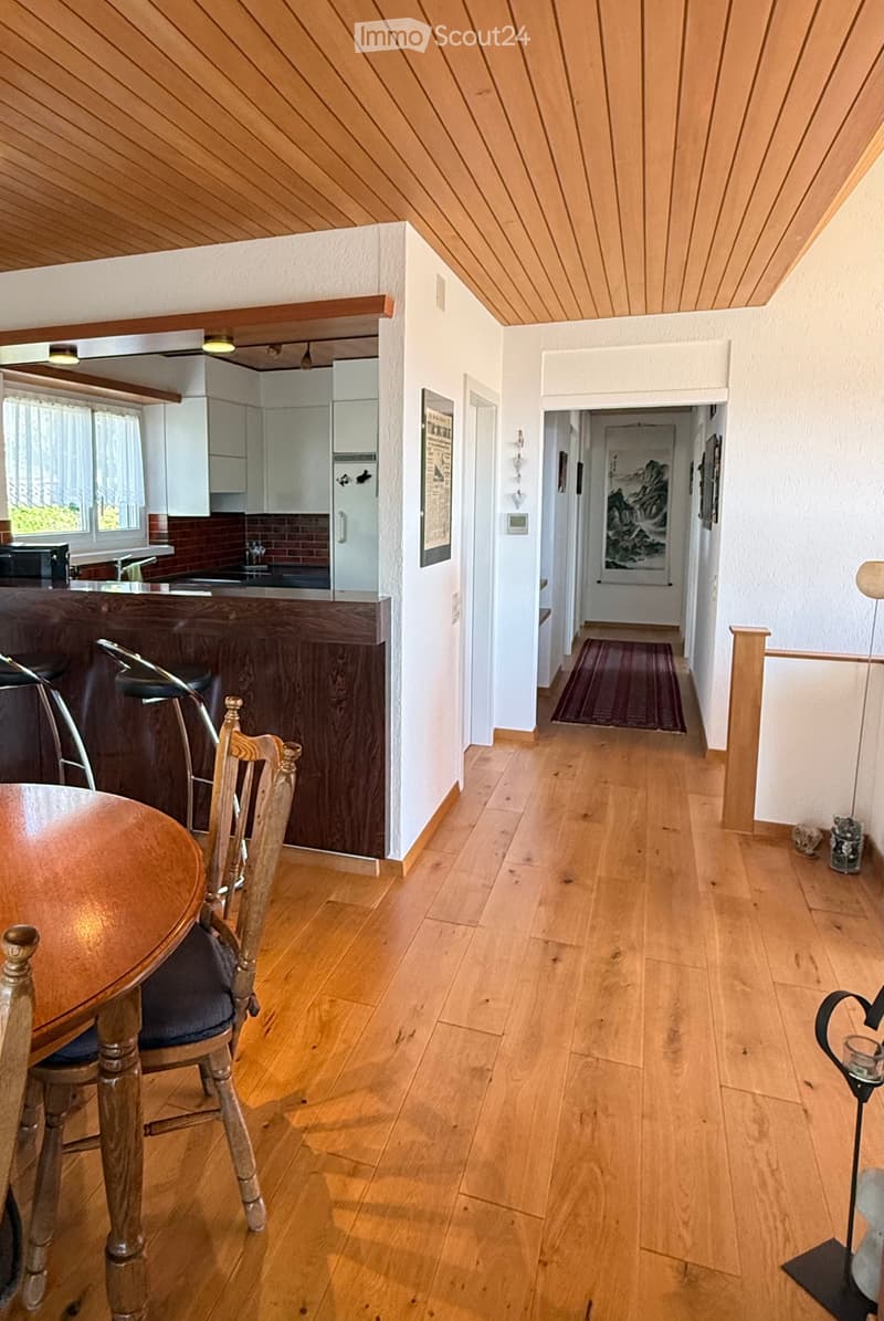 Bright hallway with wooden flooring, leading to a kitchen area with bar stools and a dining table with chairs. The kitchen features white cabinets, a countertop, and a sink.