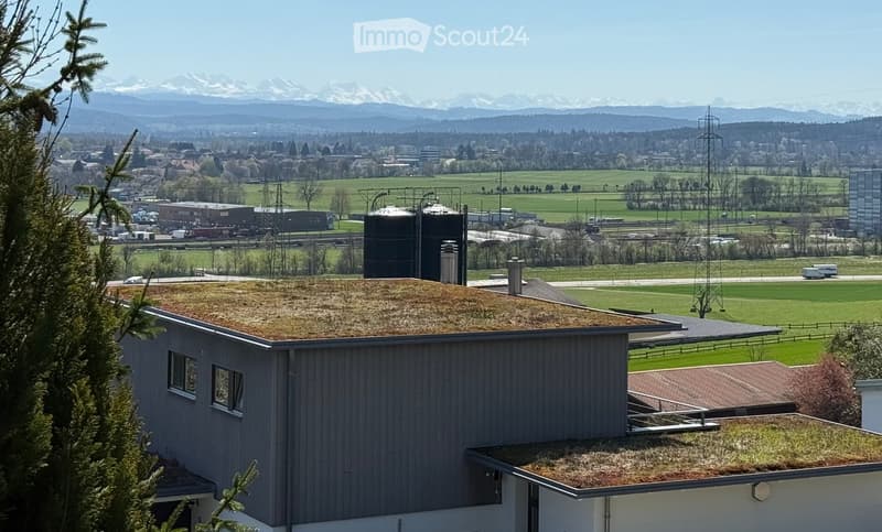 A modern building with a green roof, surrounded by open fields and distant mountains.