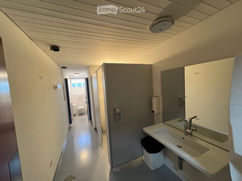 Clean, modern hallway with white walls, a double sink, and a partially visible bathroom area with a toilet and shower.