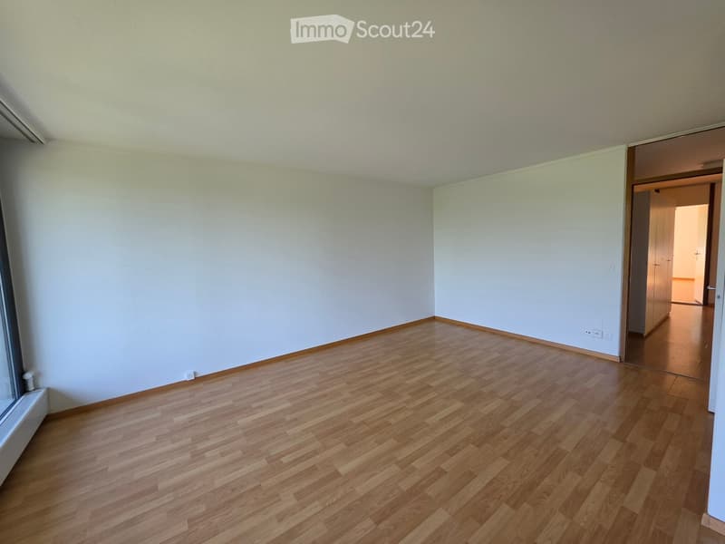Empty room with light-colored walls and wooden flooring, featuring a large window on one side and a doorway leading to another room.