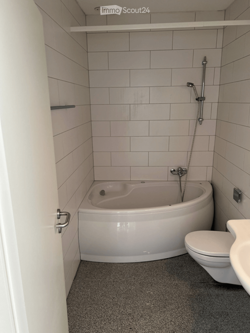 Bathroom. Modern bathroom with a corner bathtub, toilet, and showerhead. The walls are tiled in light gray, and the floor is covered with dark gray tiles. There are built-in shelves on the left wall.