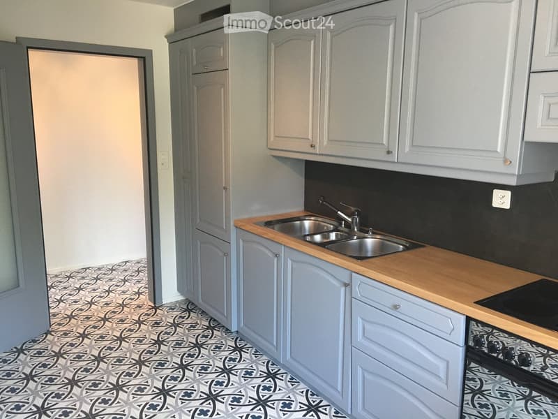 Modern kitchen with light grey cabinets, wooden countertops, stainless steel sink, and a black tiled backsplash. The floor features a patterned tile design.