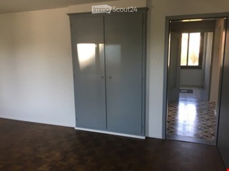 Empty room with wooden flooring, white walls, and a large grey closet. A doorway leads to another room with a tiled floor and a window.