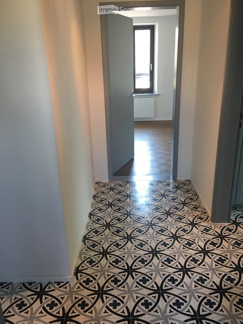 Hallway with patterned tile floor, light-colored walls, and an open door leading to another room with wooden flooring.