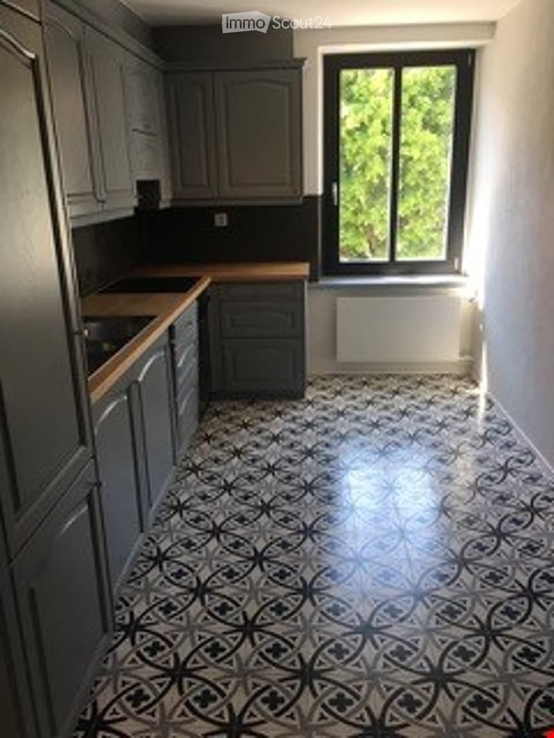 Modern kitchen with grey cabinets, wooden countertops, and a distinctive black and white patterned tile floor. A large window allows natural light to fill the space.