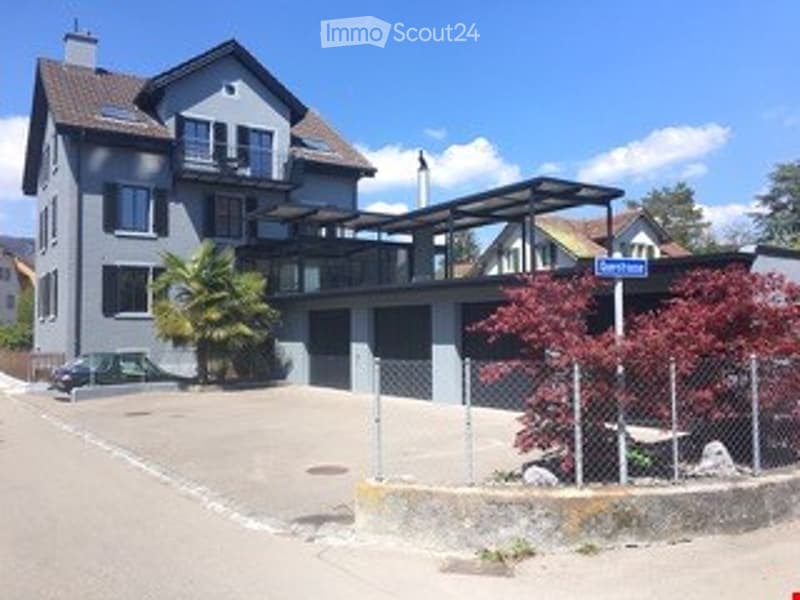 Modern three-story house with grey facade, multiple balconies, garage, and parking area.