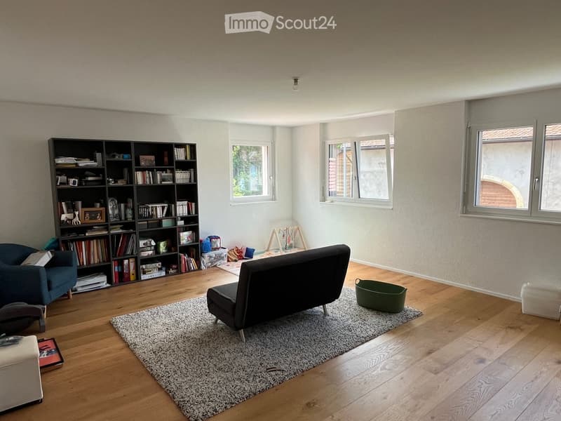 Modern living room with wooden flooring, large windows, a dark grey sofa, a blue armchair, a bookshelf filled with books, and a small rug.