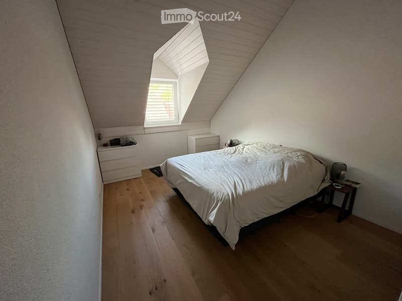 Bedroom with wooden flooring, a bed with white bedding, a nightstand, and a drawer unit. The room has slanted ceilings and a skylight window.