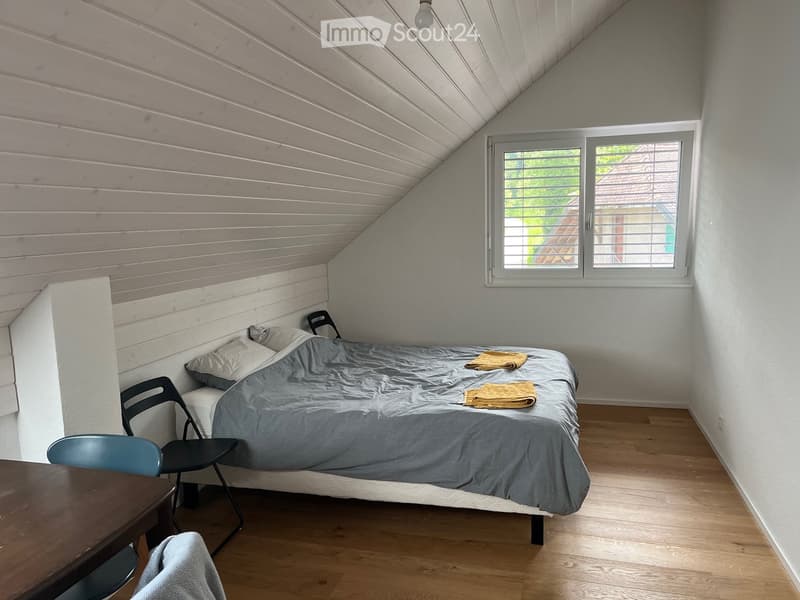 Cozy attic bedroom with wooden slat ceiling, grey bedding, two chairs, and a small table.