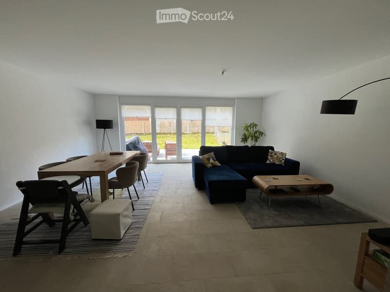 Modern living room with a blue sectional sofa, coffee table, ottoman, dining table with chairs, and large windows leading to a patio.