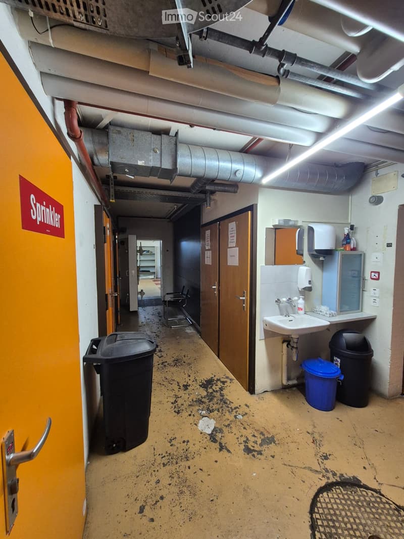 Corridor with yellow walls, pipes on ceiling, sprinkler sign, trash bins, sink, chair, steel shelves