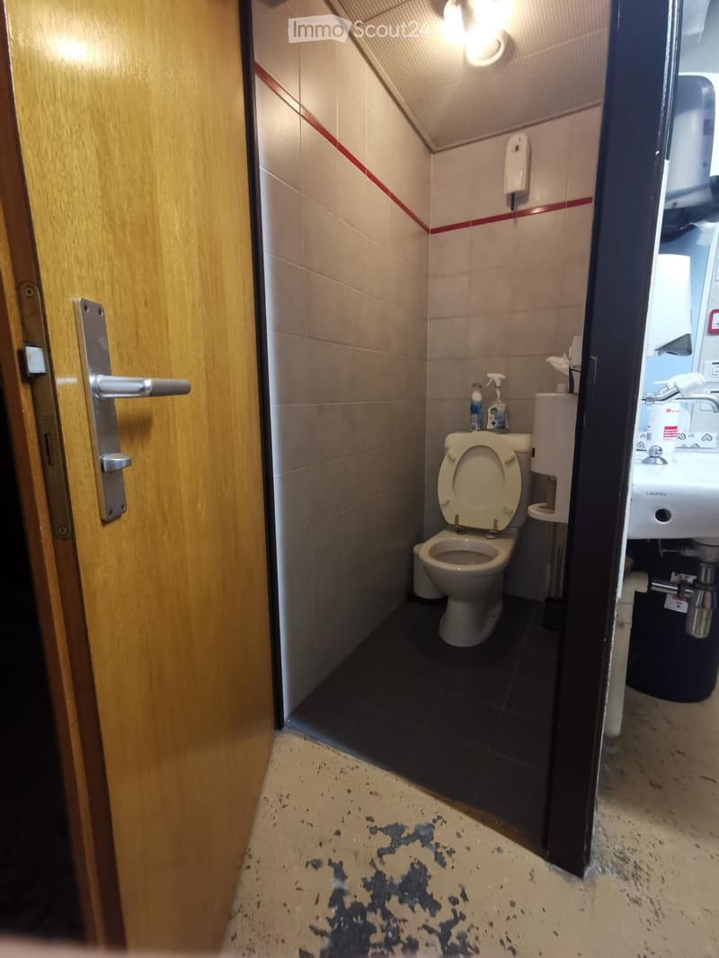 Small bathroom with a toilet, sink, and hand sanitizer. The floor has visible stains, and the walls are tiled with a red accent line.