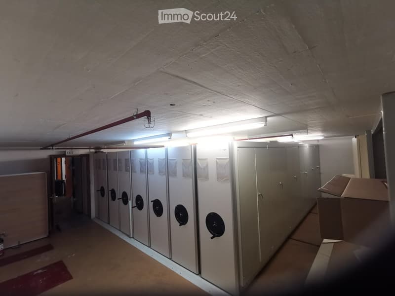 A narrow, industrial-style hallway with white lockers lining one wall, fluorescent lighting, and a visible 'EXIT' sign. The floor is concrete, and the walls are painted white.