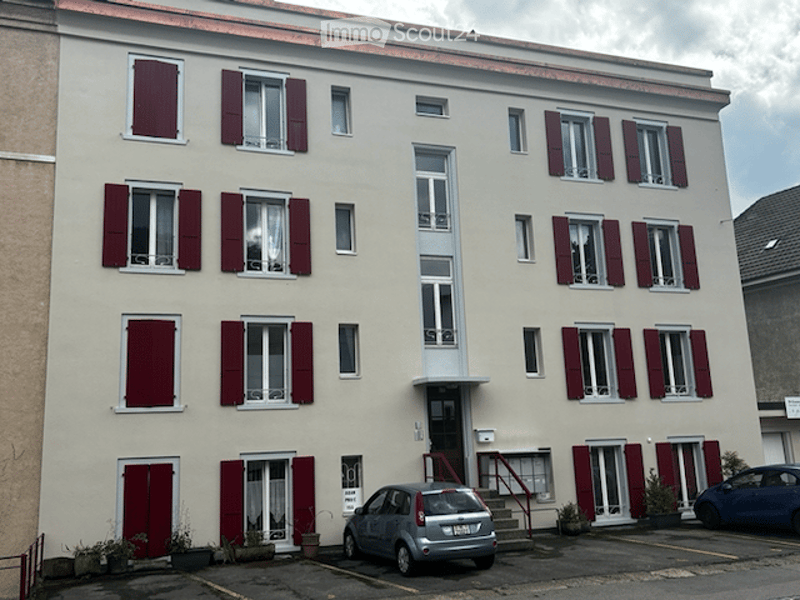 Four-story residential building with beige facade, red shutters, and multiple windows. A small parking area is visible in front with a few cars parked.