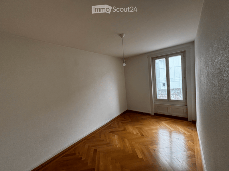 Empty room with wooden herringbone flooring, white walls, and a single window with white frame and glass panes. A single ceiling light fixture is visible.