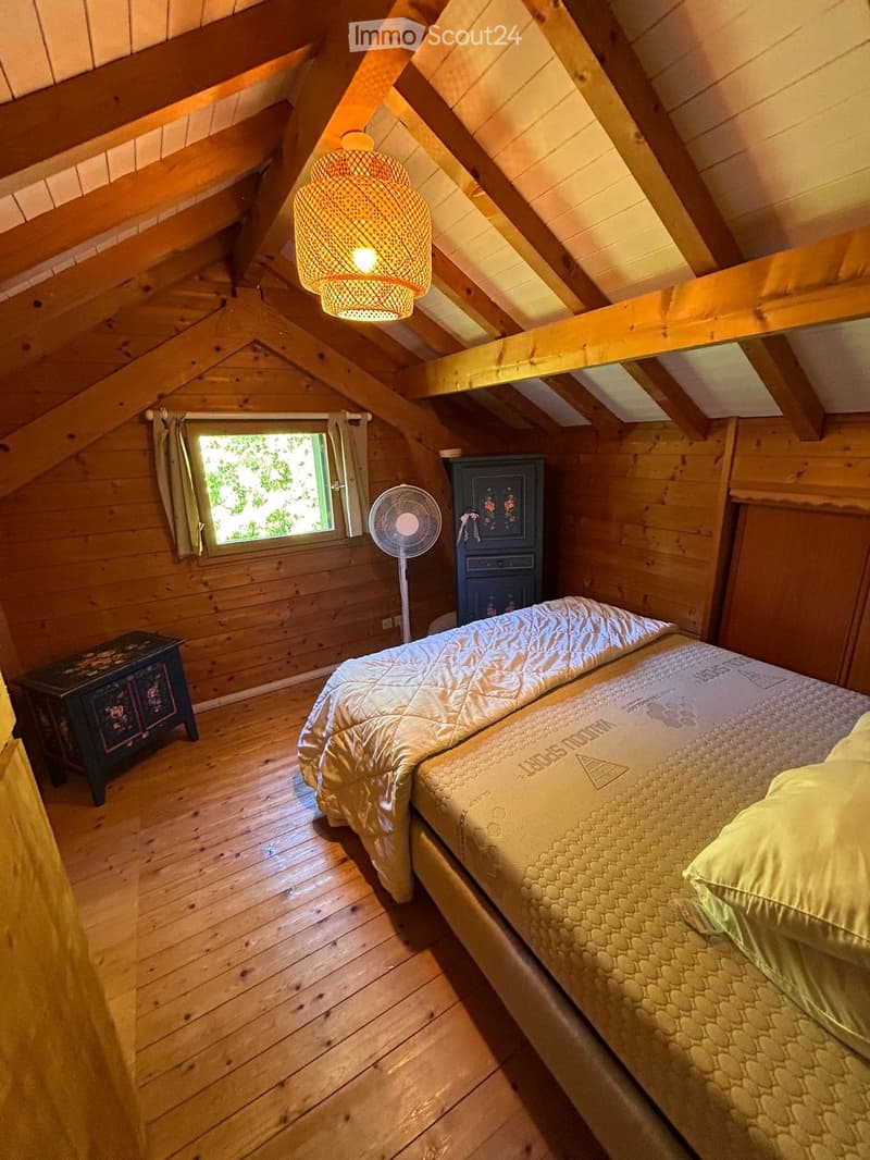 Cozy attic bedroom with wooden beams, slanted ceiling, and exposed wooden floor. Features a single bed with a quilt and pillows, a small nightstand, a standing fan, and a wicker pendant light.
