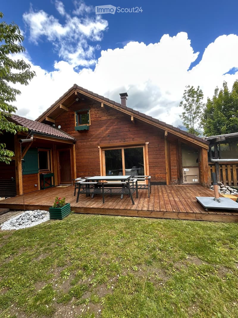 A wooden house with a pitched roof, large windows, and a wooden deck with outdoor furniture and a small garden area.