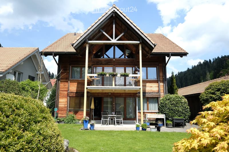 House. Two-story wooden house with a triangular roof, large windows, and a balcony. The house is surrounded by a well-maintained garden with a patio area, outdoor furniture, and potted plants.