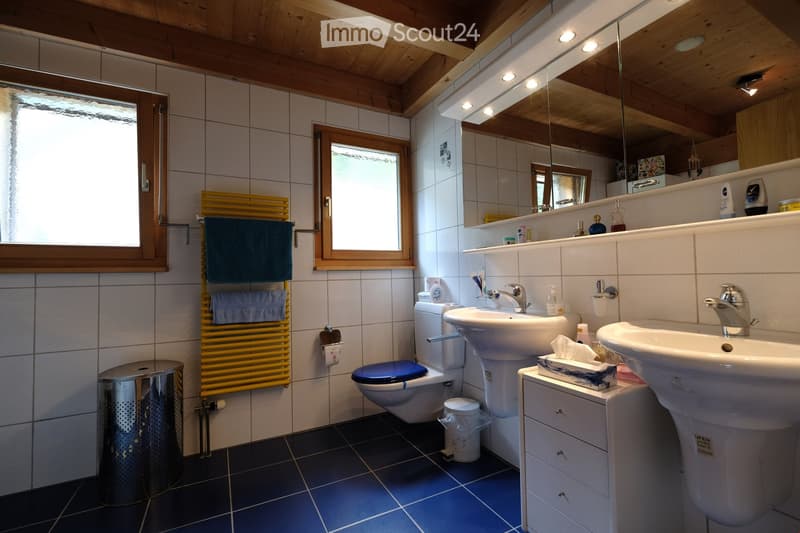 Bathroom. Bathroom with two sinks, a toilet, a large mirror, a towel rack with towels, and a trash can. The room features wooden beams on the ceiling and tiled walls and floors.