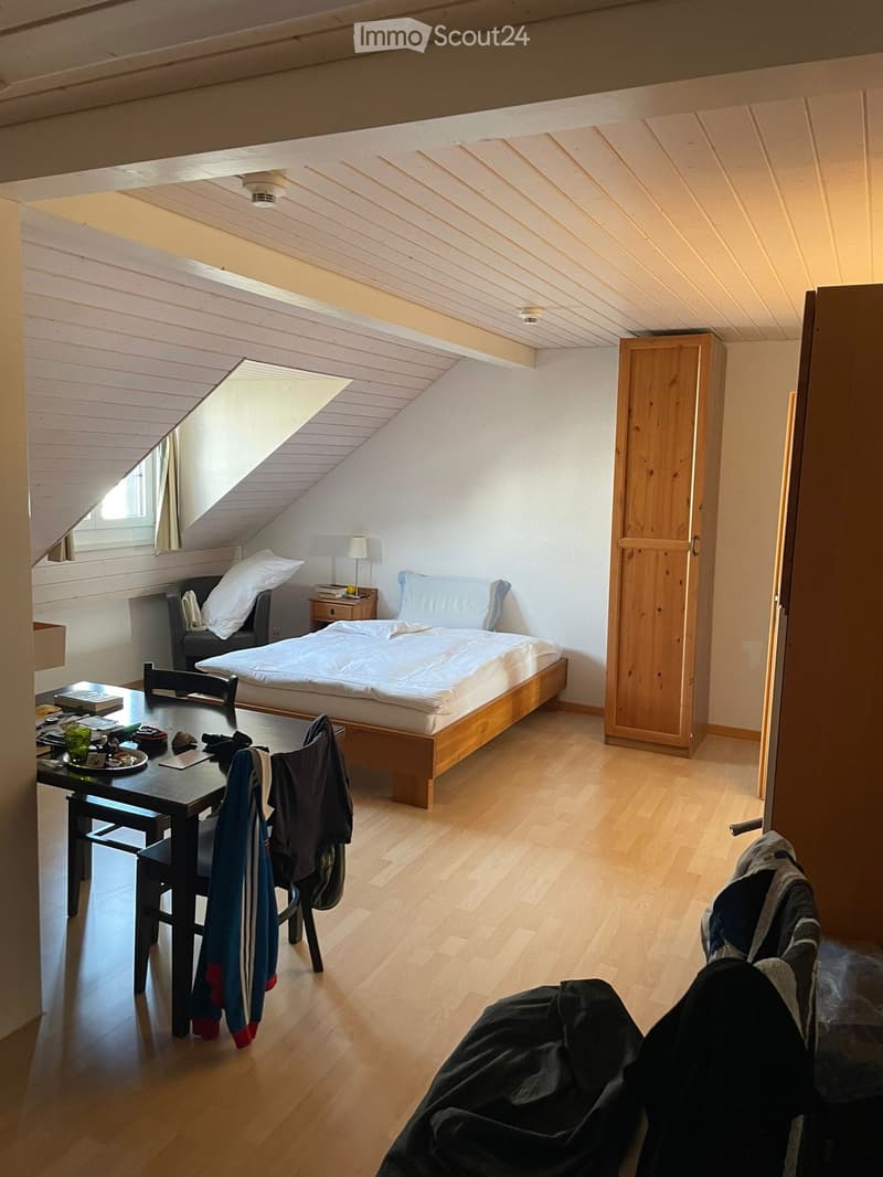 Room with wooden floor, white walls, bed with pillows, desk with chairs, wooden cabinet, table with plates and books, table lamp, and an attic window with curtains.