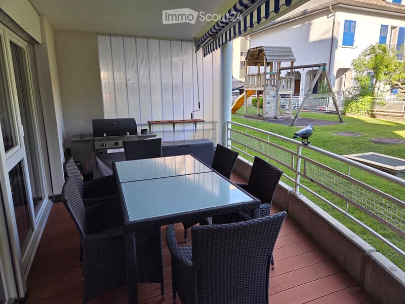 Balcony with a table, chairs, and a grill, overlooking a grassy area with a playground.