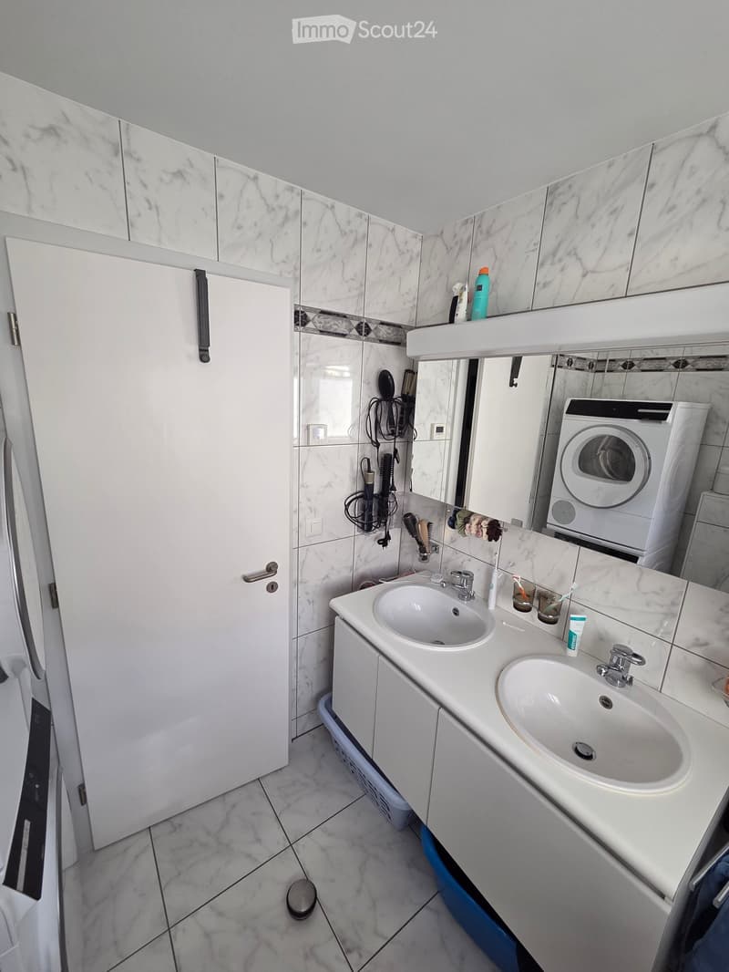Modern bathroom with white marble tiles, double sink vanity, large mirror, and a washing machine in the corner.