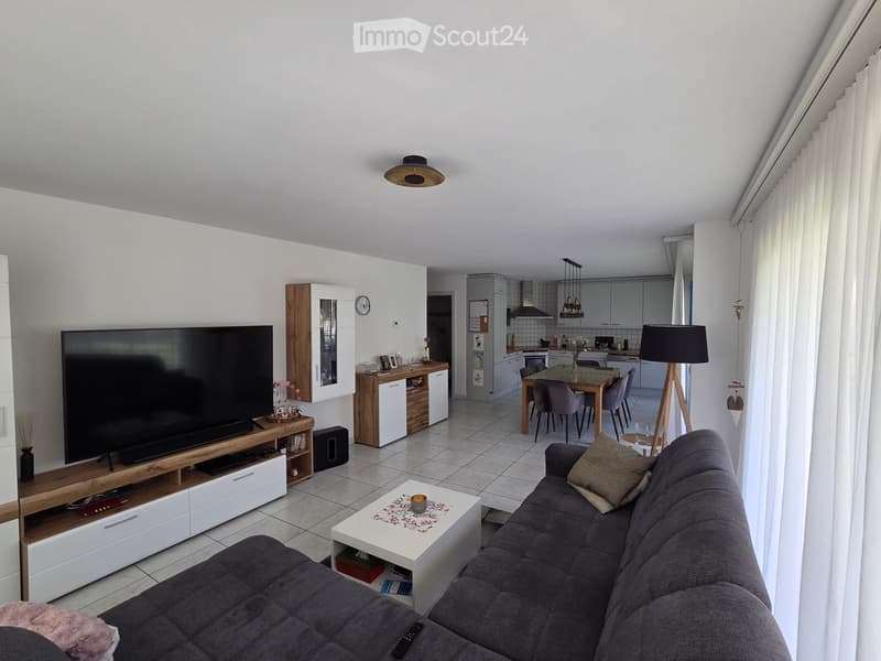 Open-plan living and dining area with a grey sofa, coffee table, TV, dining table with chairs, and a modern kitchen in the background.