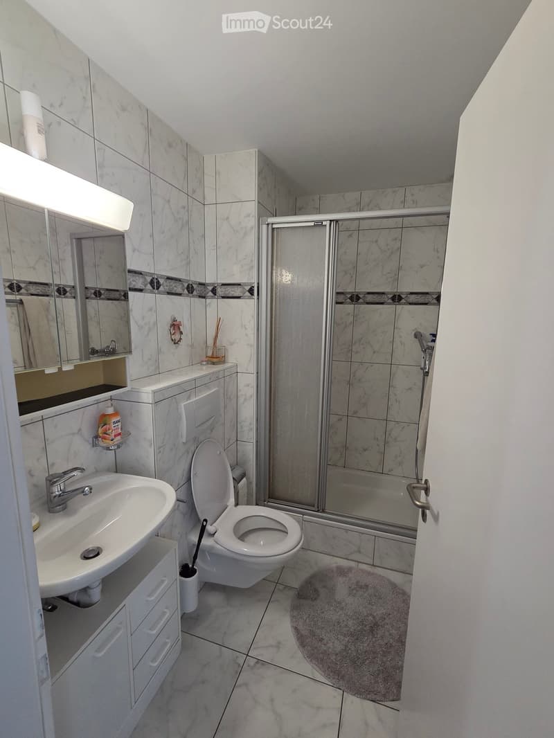 Modern bathroom with white marble tiles, a sink with a chrome faucet, a toilet, a shower with a glass door, a mirror with built-in lighting, and a small bathroom rug on the floor.