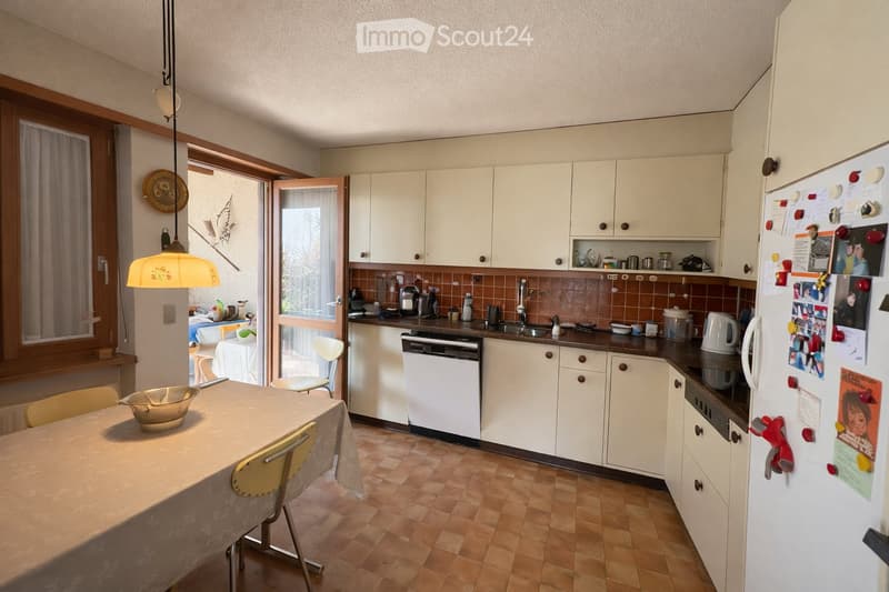 A well-equipped kitchen with white cabinets, a tiled backsplash, and a central island. The kitchen features a refrigerator, dishwasher, sink, and various appliances. A dining area with a table and chairs is visible, along with a hanging light fixture and