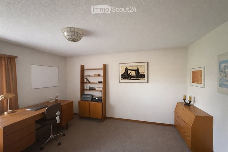 A well-lit, furnished room with a wooden desk, office chair, lamp, bookshelf, printer, and decorative items including candles and artwork.