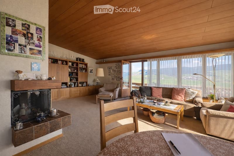 Bright living room with wooden ceiling, large windows offering a scenic view, fireplace, sofa, coffee table, armchair, bookshelf, and a round table with a notebook and pen.