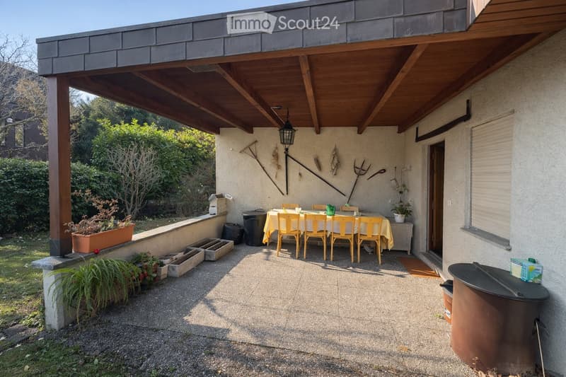Outdoor patio with a wooden ceiling, a dining table with yellow chairs, potted plants, and garden tools on the wall.