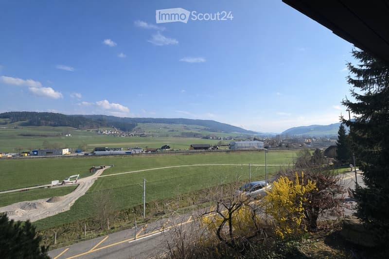 A panoramic view of a rural landscape with green fields, scattered buildings, and distant hills under a clear blue sky.
