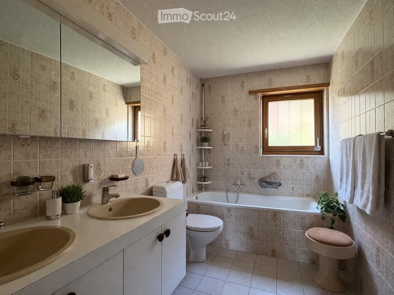 Bathroom with beige tiles, a sink, toilet, bathtub, and a window. Includes a mirror, towel, and small plant on the counter.