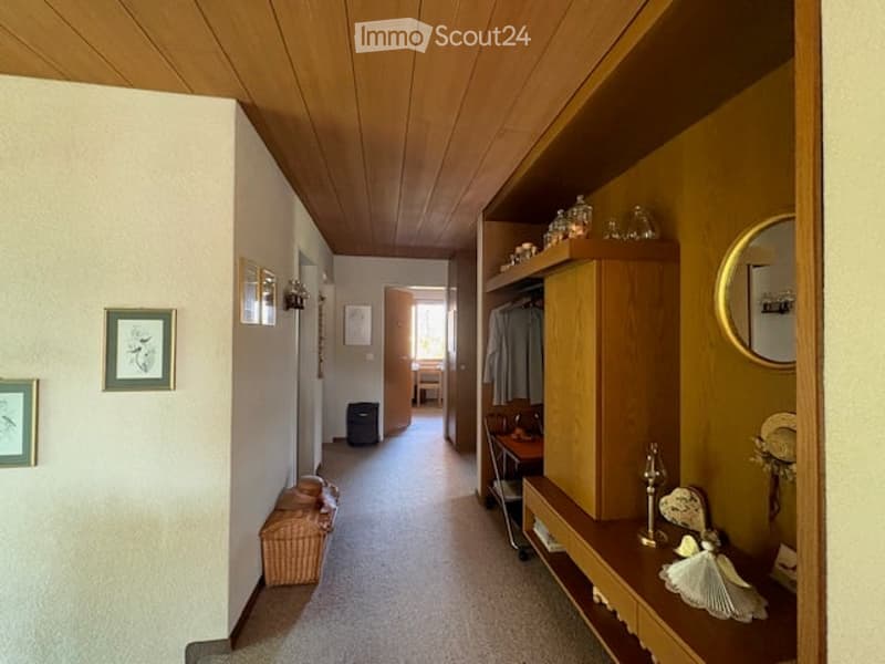 A well-lit hallway with wooden ceiling, neutral-colored walls, and a carpeted floor. Features include built-in wooden shelving, framed artwork, and a small wicker basket on the floor.