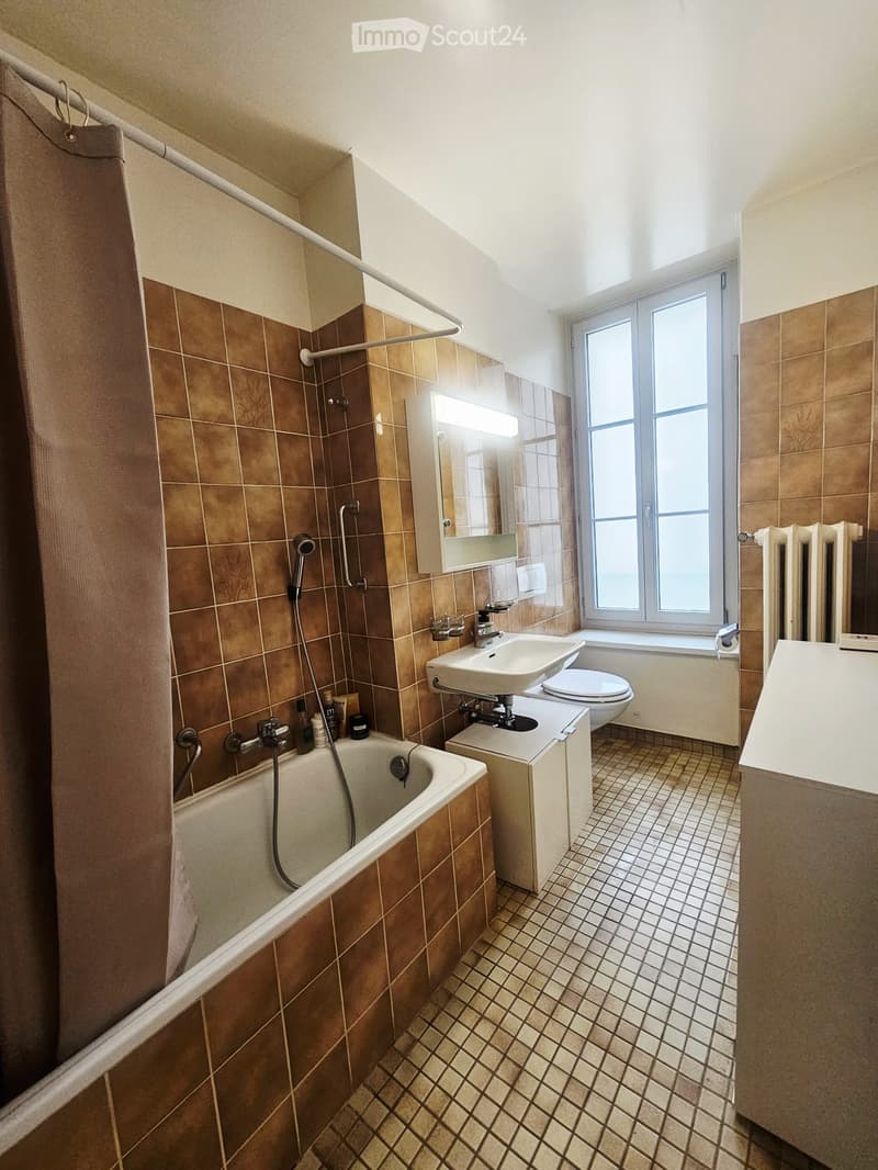 Bathroom with brown tiled walls and floor, bathtub, toilet, sink, mirror, radiator, and window.
