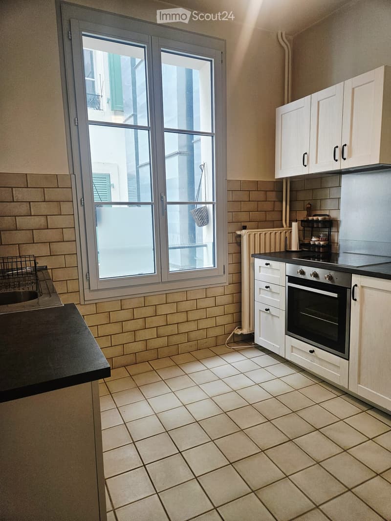 Modern kitchen with tiled walls and floor, white cabinets, black countertop, and stainless steel oven. Features a large window with a view of the street.