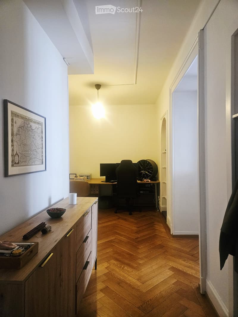 A furnished room with a wooden herringbone floor, a desk with a computer and chair, a cabinet with a cup and bowl, and a picture frame on the wall.