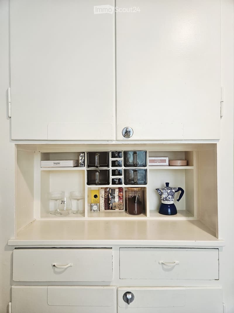 Open kitchen cabinet with glassware, books, and a coffee pot displayed on shelves.