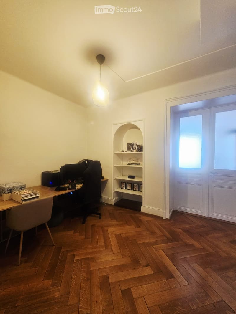 A well-lit study or office space with wooden flooring, a large wooden desk, a comfortable chair, a computer setup, a hanging lamp, and a built-in arched shelf with picture frames and books.