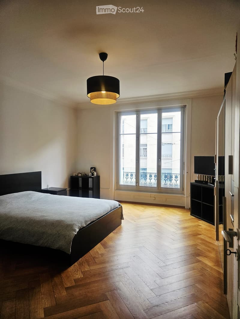 Modern bedroom with wooden flooring, large window with balcony access, bed with dark headboard, nightstand, cabinet, and television.