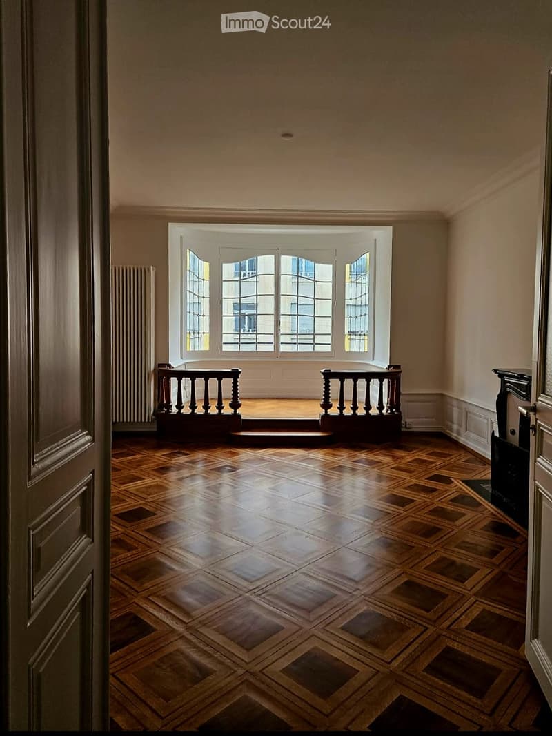 An empty room with wooden herringbone flooring, large windows with white frames, and a small balcony area with a wooden railing.