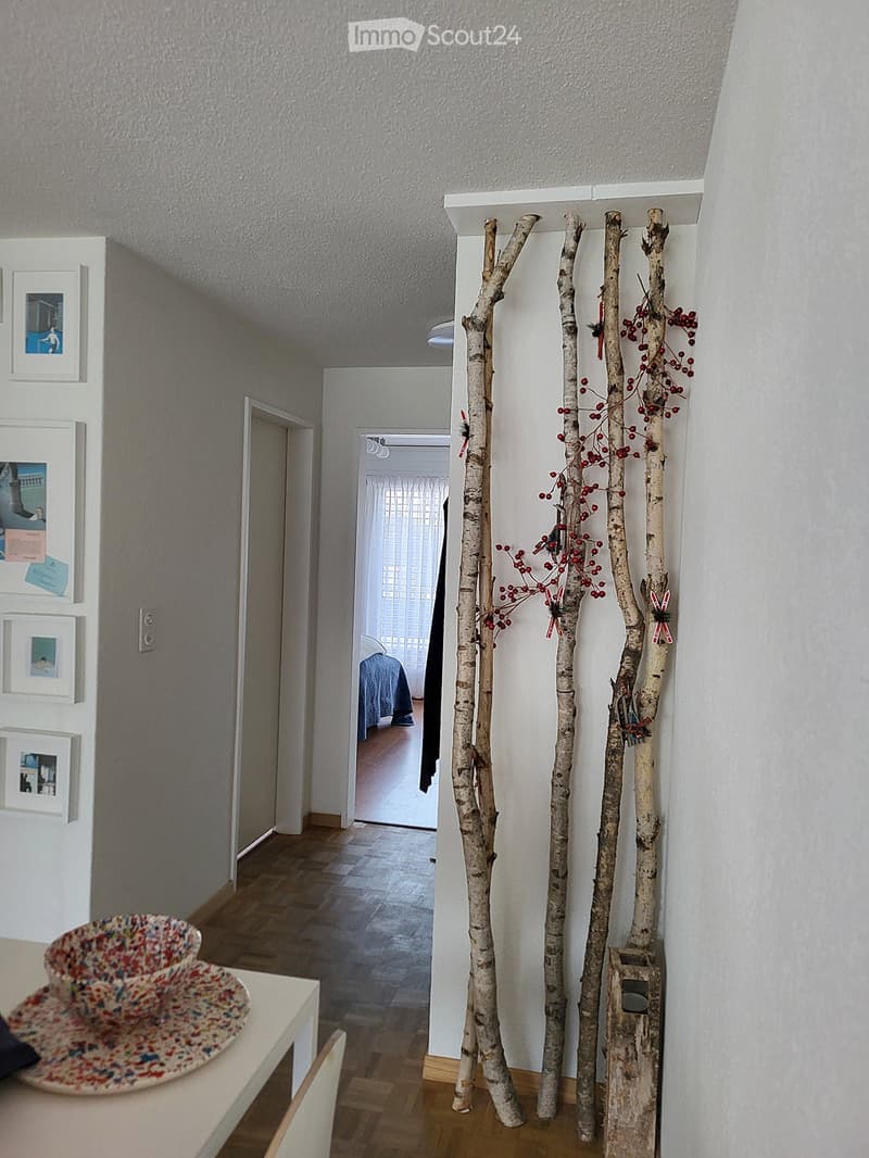 A narrow hallway with wooden flooring, white walls, and several framed pictures on the left wall. A decorative arrangement of birch branches with red berries is placed on the right side. A small table with a colorful bowl is visible on the left side near